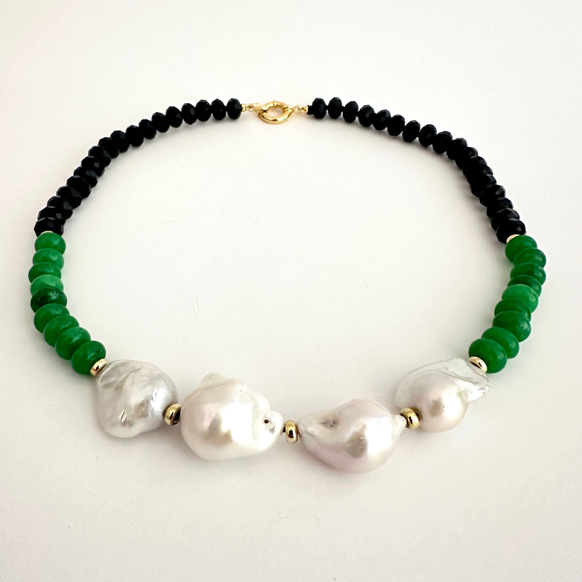 A beaded necklace featuring black round beads, vibrant green faceted beads, and four large white baroque pearls, accented with small gold spacers and a gold clasp, displayed on a clean white background.