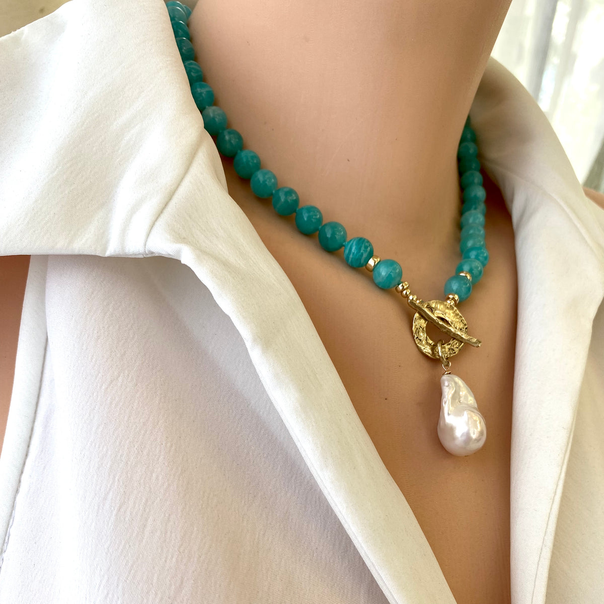 A person wearing a white shirt showcasing a blue amazonite and white baroque pearl necklace with gold-filled accents and a gold bronze toggle clasp.