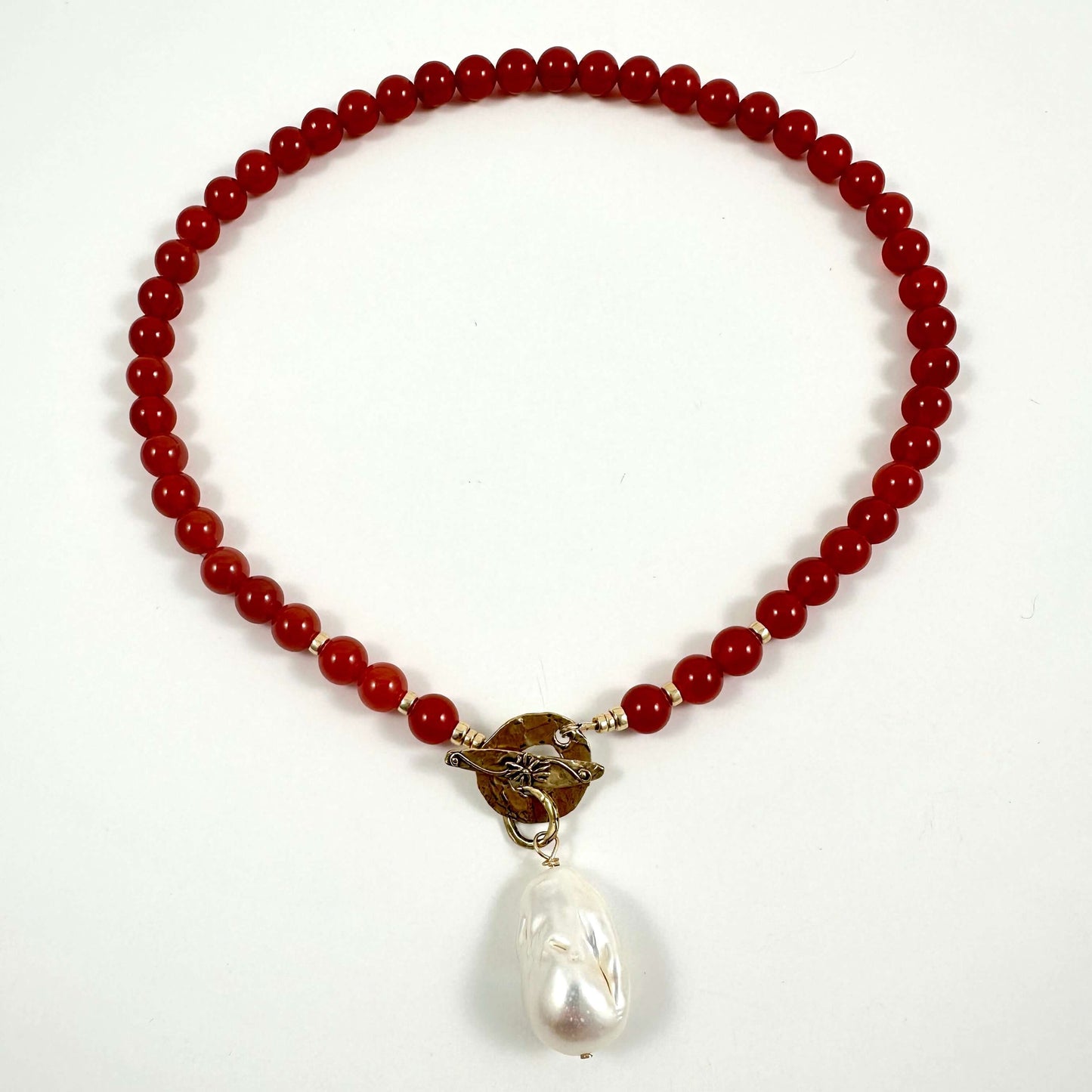 A beaded necklace featuring deep red round beads, a brown textured central accent, and a dangling white baroque pearl, displayed on a clean white background.