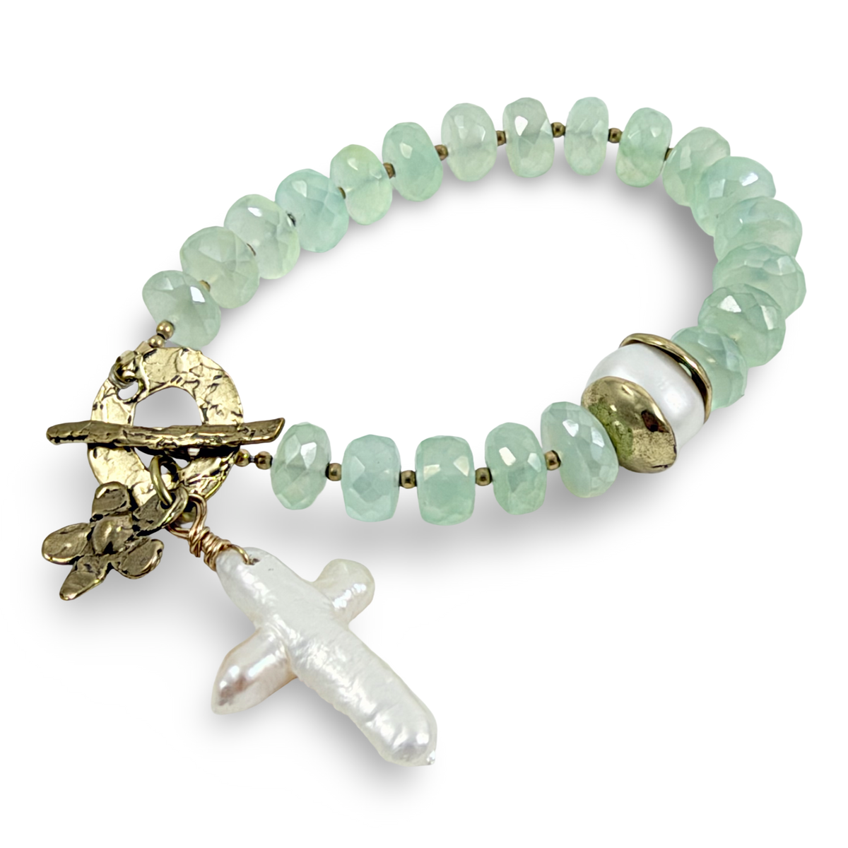 A beaded stretch bracelet featuring light green faceted beads, a textured gold-tone toggle clasp with a floral charm, a white pearl baroque cross pendant, and a white and gold spacer bead, shown on a white background.