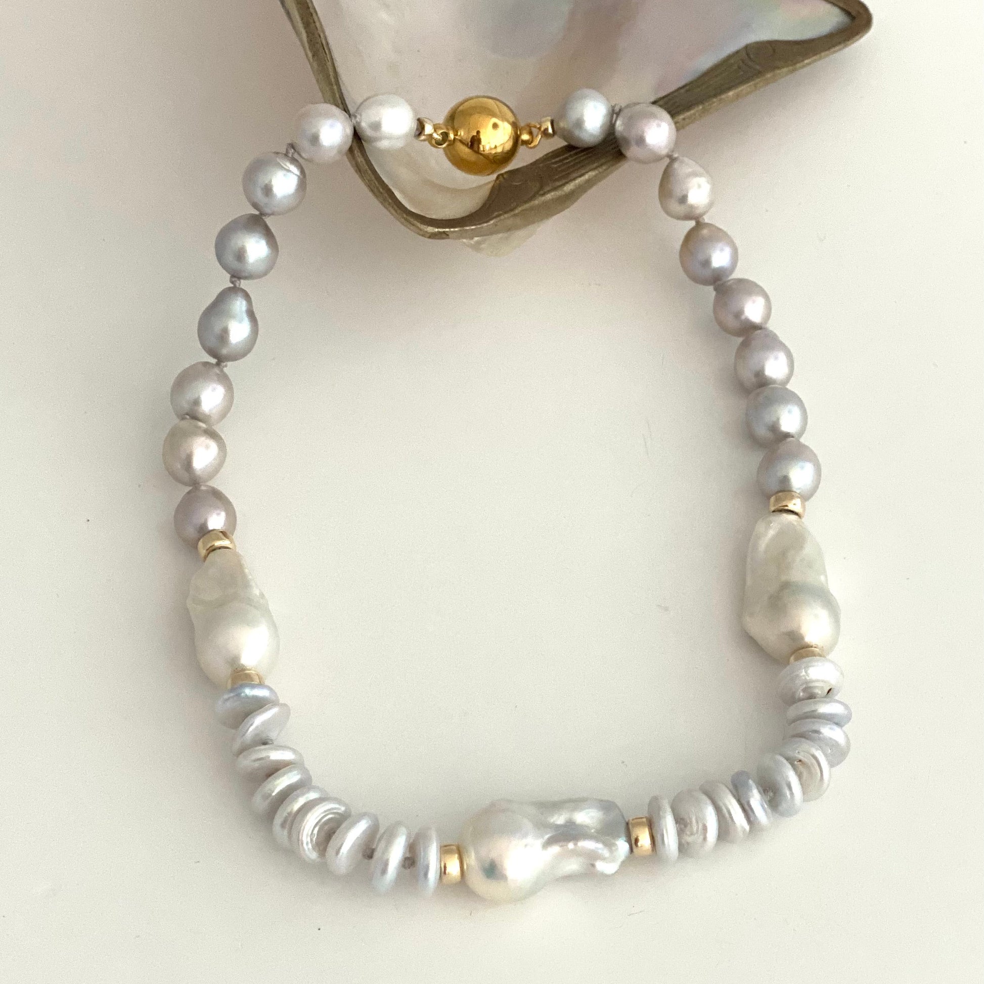 HAND KNOTTED NECKLACE made of white and silver baroque pearls, silver PEARL spacer beads, and gold bead, displayed on a white surface with a piece of mother-of-pearl shell at the top.
