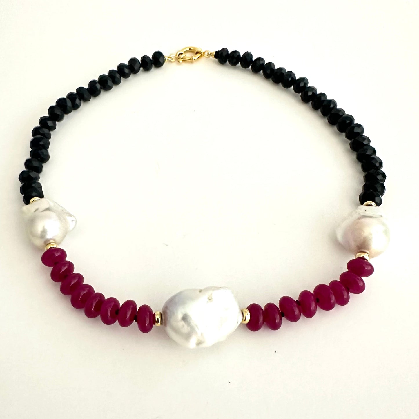 A beaded necklace featuring black faceted beads, deep red faceted beads, and three white baroque pearls, accented with small gold spacers and a gold clasp, shown on a plain white background.