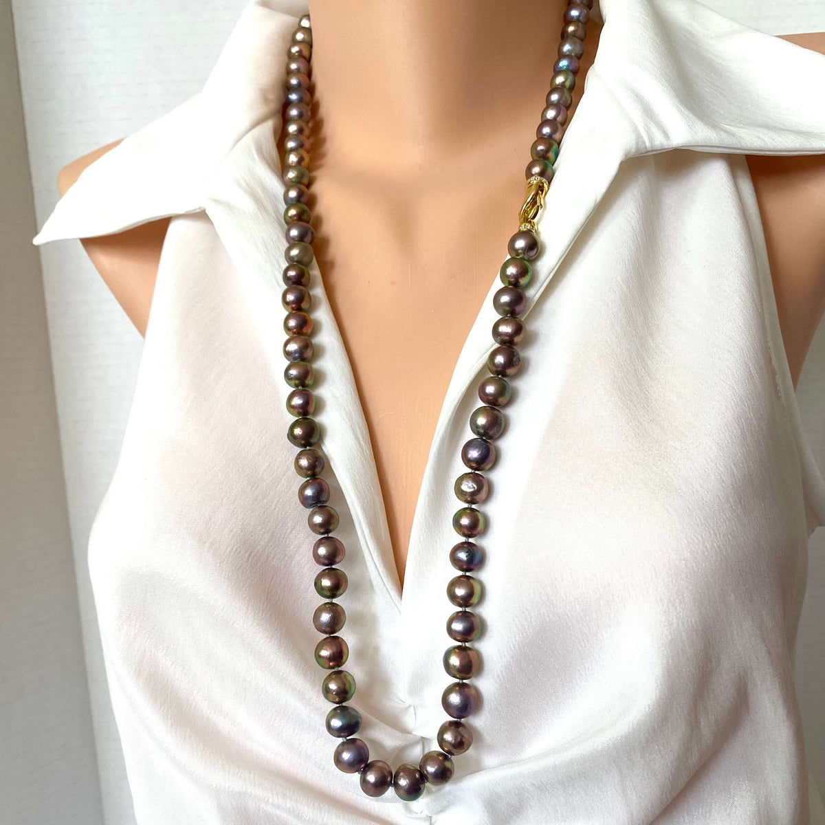 A long strand of dark, iridescent freshwater pearls with a gold clasp, displayed on a mannequin torso wearing a white sleeveless blouse.