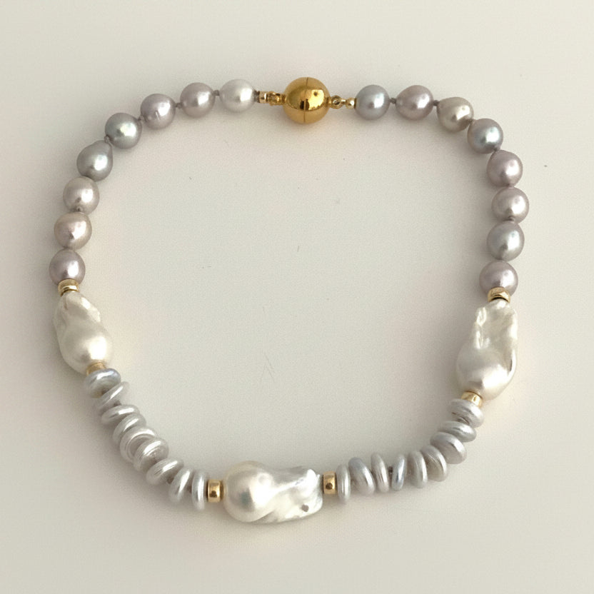 A beaded bracelet featuring a strand of small gray pearls, a few larger white baroque pearls, textured gray beads, and gold spacer accents, displayed on a light beige background.