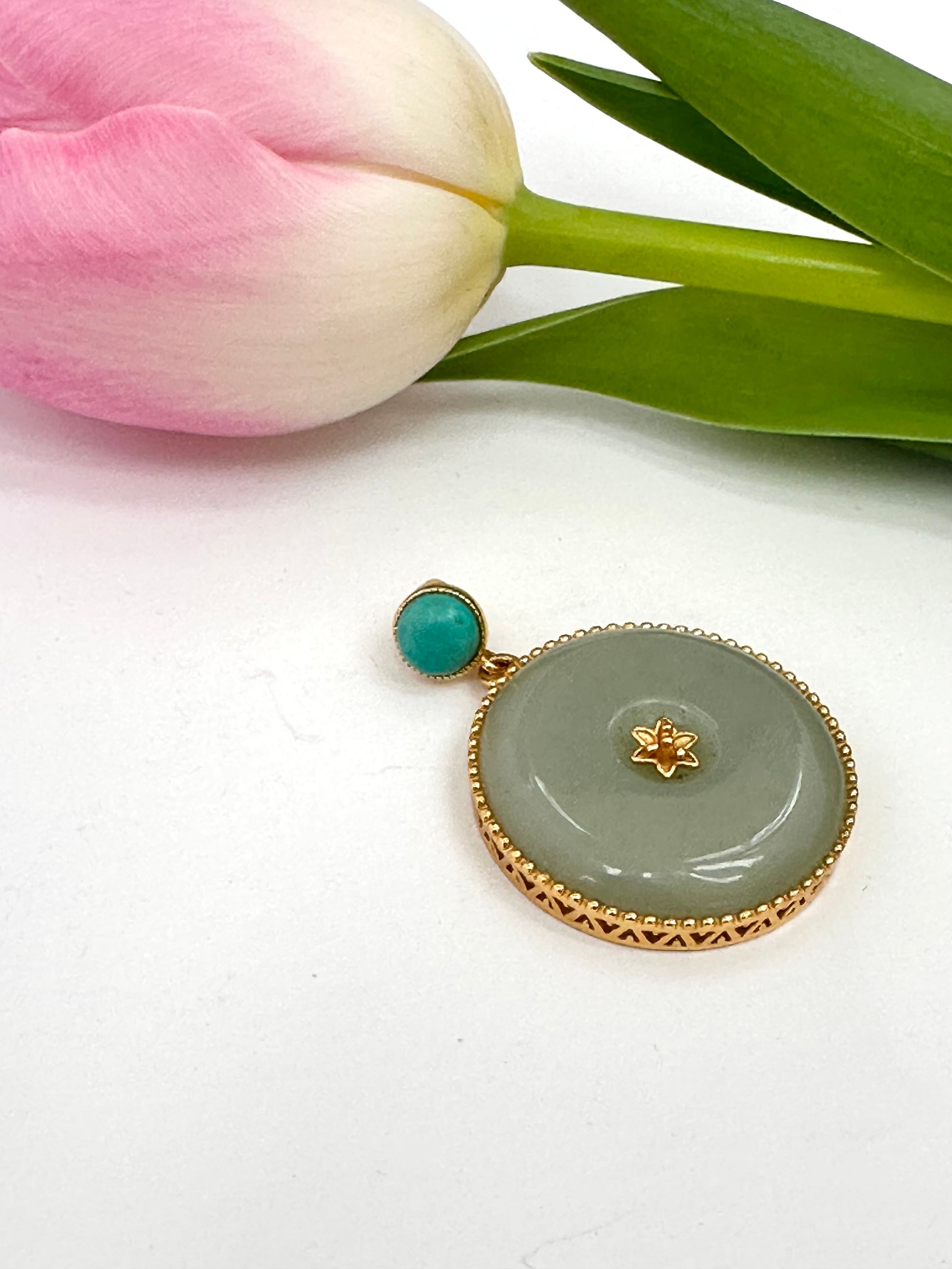 Jade pendant with gold accents and turquoise stone on a white surface with a pink tulip.