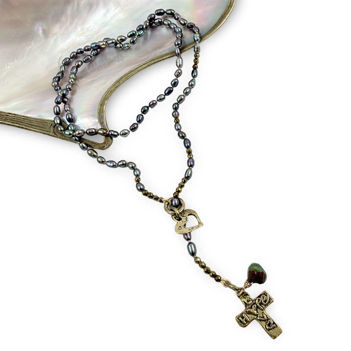 A beaded rosary necklace with dark gray and black pearls, antique bronze-toned metal links, a small heart-shaped charm, and a cross pendant, displayed on a white background with a curved iridescent shell prop.