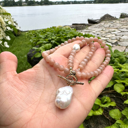Sunstone necklace with baroque pearl pendant held in hand against green foliage