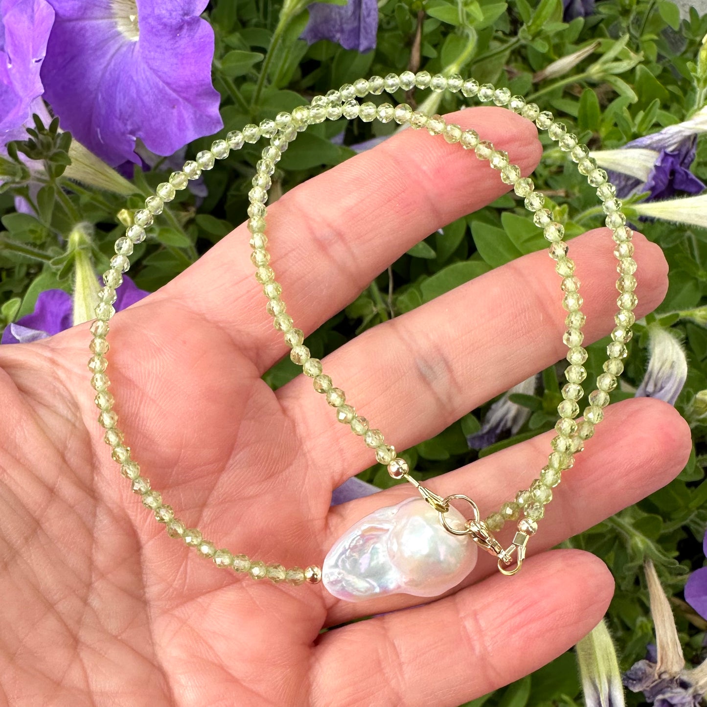 Peridot and baroque pearl necklace held in hand against purple flowers