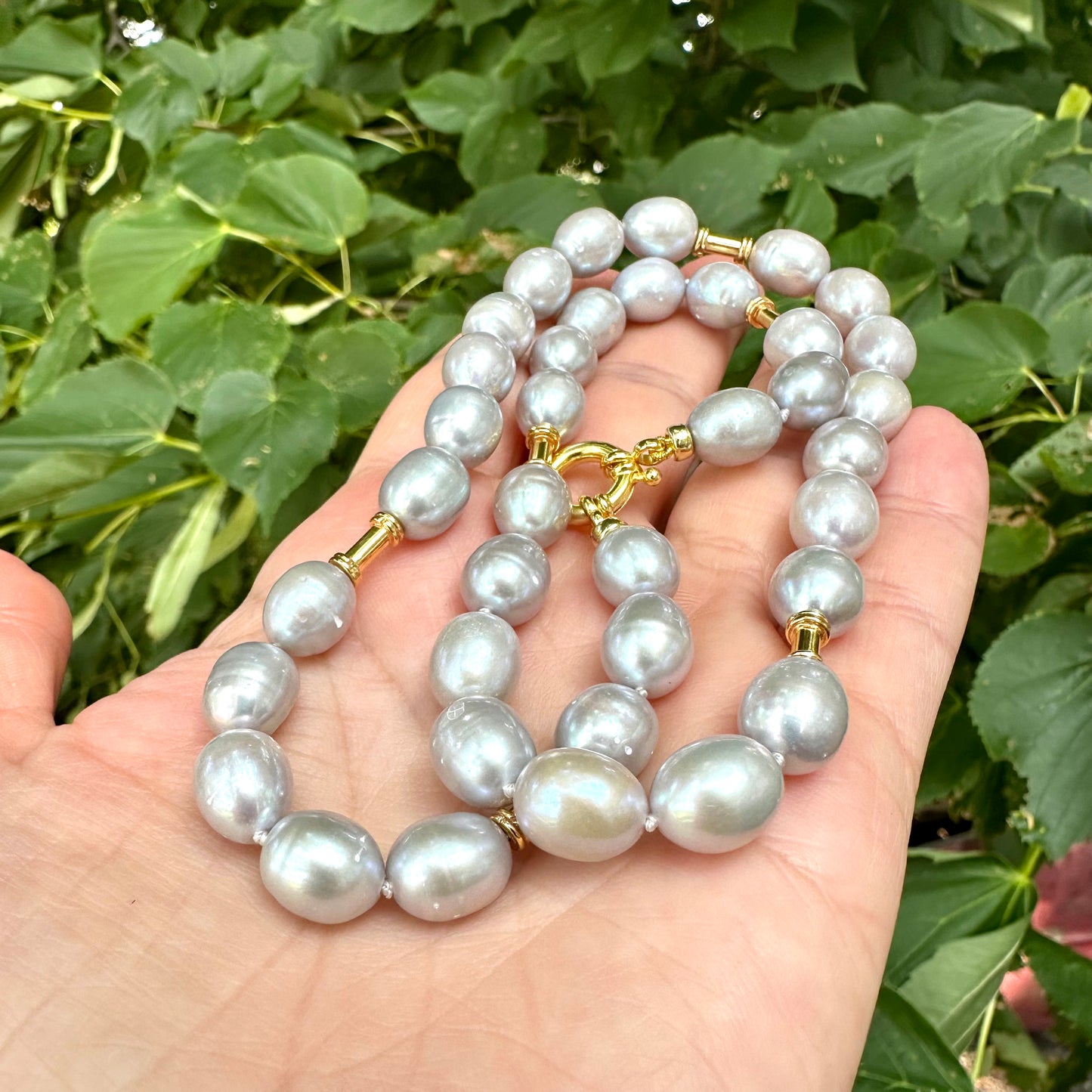 Grey pearl necklace with gold details held in hand against green foliage