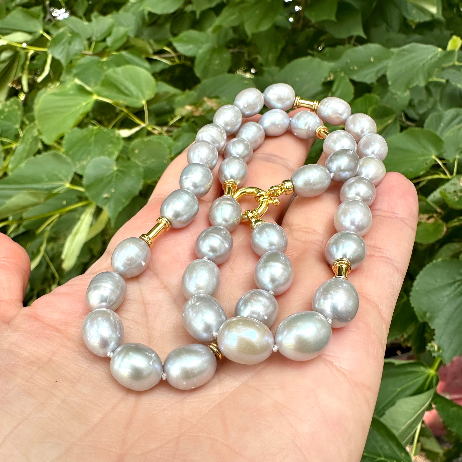 Grey pearl necklace with gold details held in hand against green foliage