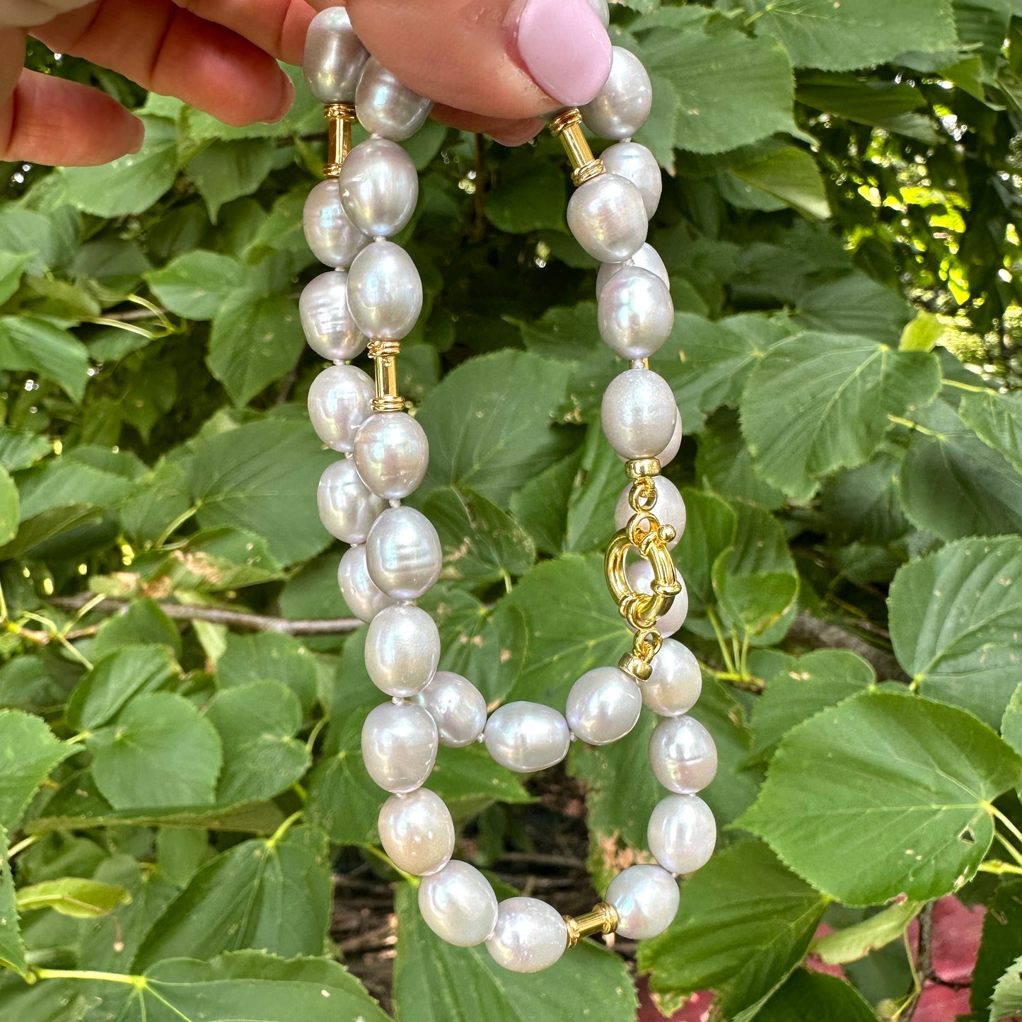 Close-up of grey pearl necklace with gold details held in hand near green leaves