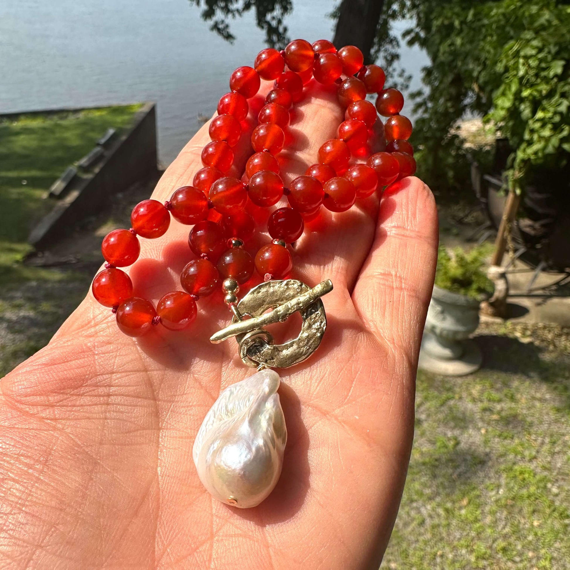 Close-up of red agate toggle necklace held in hand with lake and greenery background