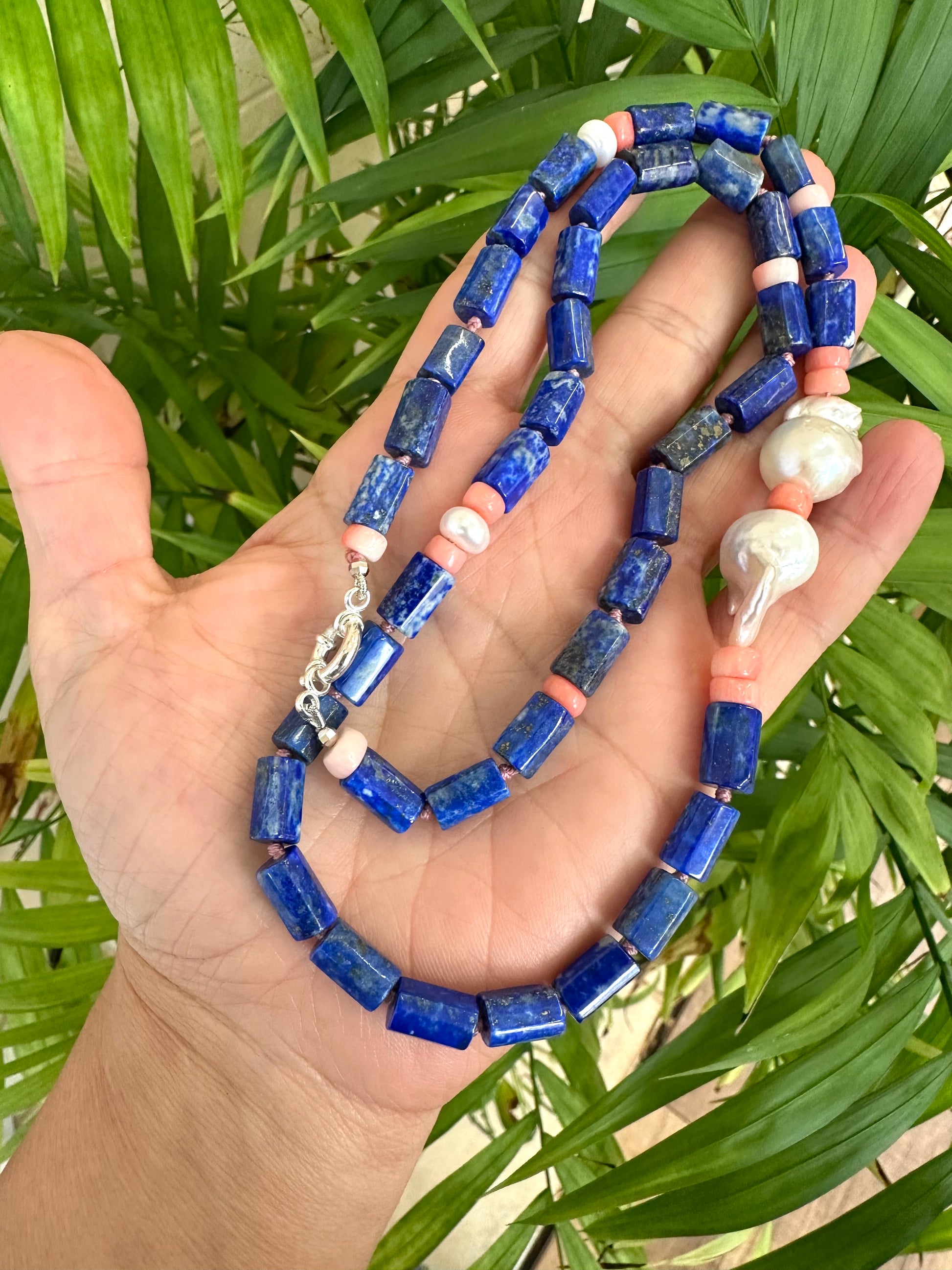 Lapis lazuli necklace with pink coral and baroque pearls held in hand, showcasing vibrant blue, pink, and pearl elements.