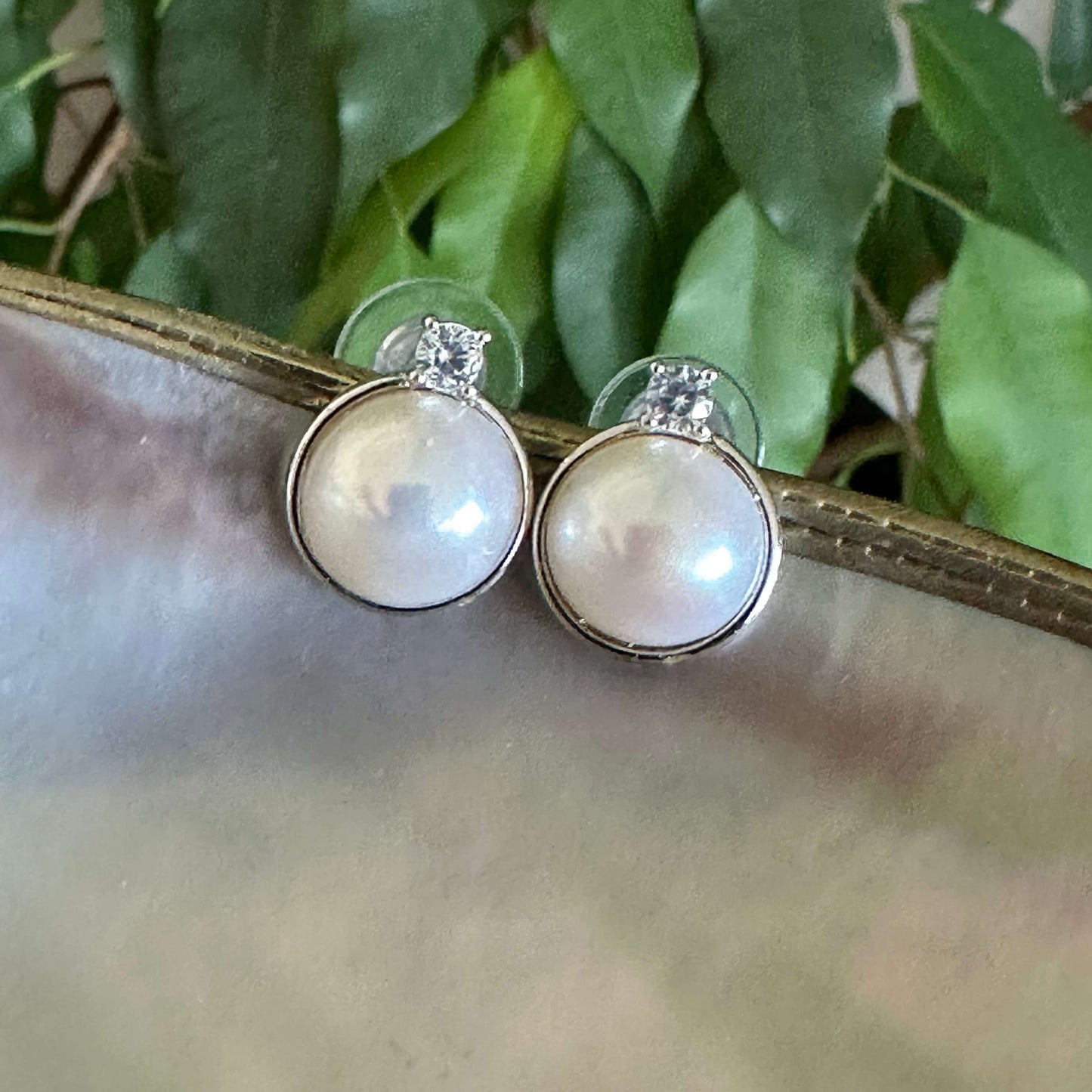 White mabe pearl stud earrings with CZ on white shell and green leaves, reflecting natural light and silver-white radiance.