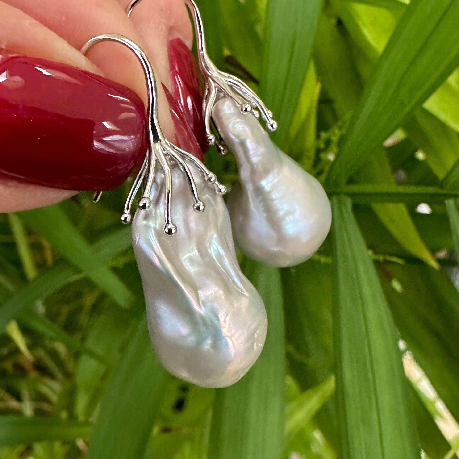Silver grey baroque pearl drop earrings held in hand against green leaf backdrop, showing natural light reflection and scale.