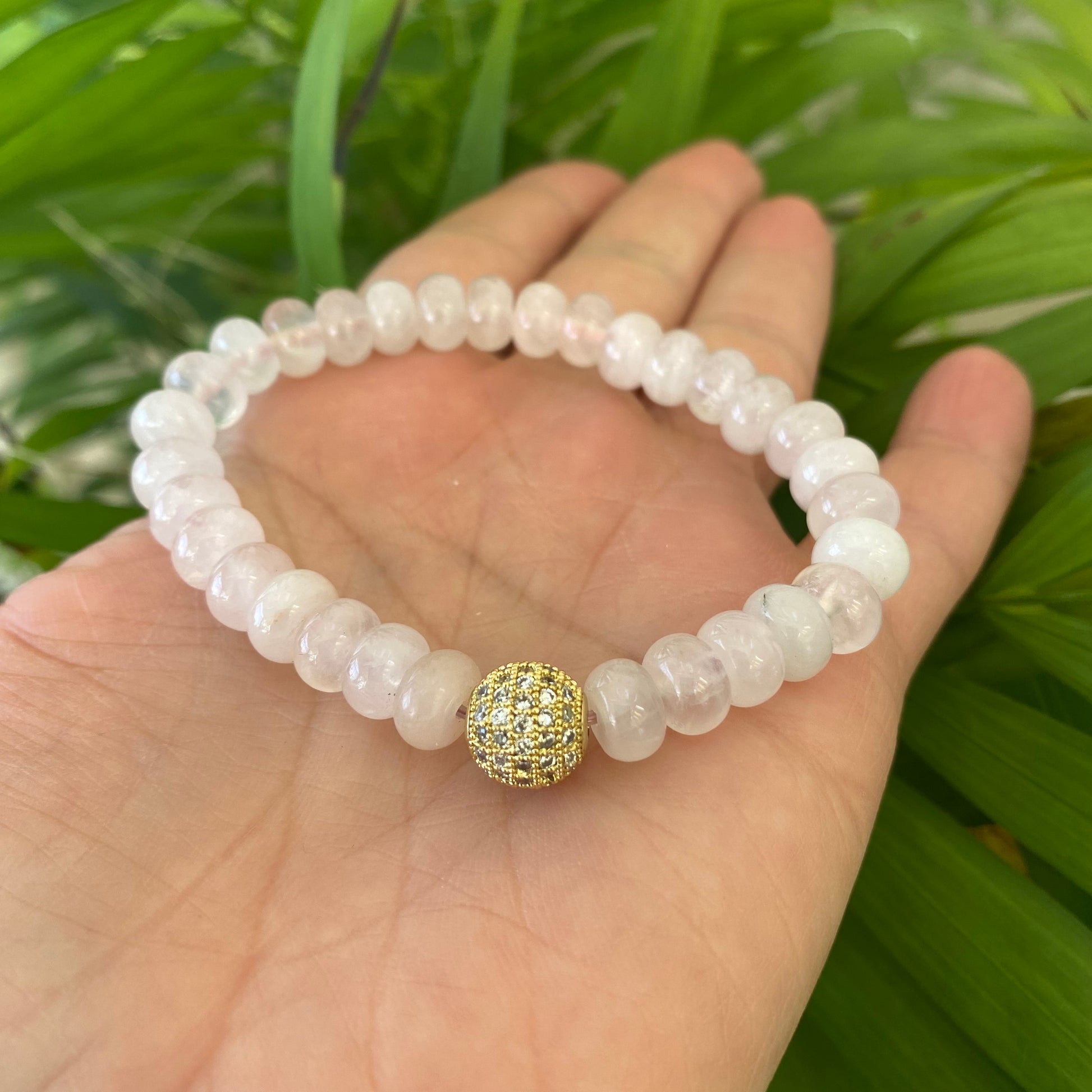 Rose quartz stretch bracelet with gold accent bead displayed on a hand surrounded by greenery.