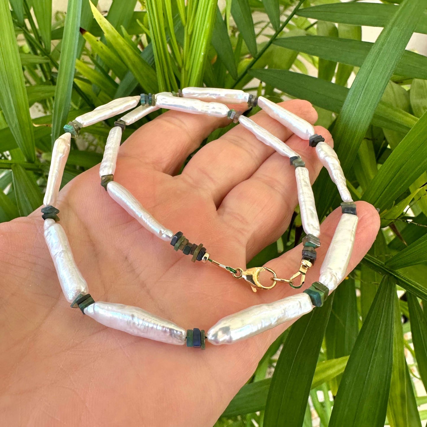 Hand holding biwa pearl and black opal necklace against green leaf backdrop