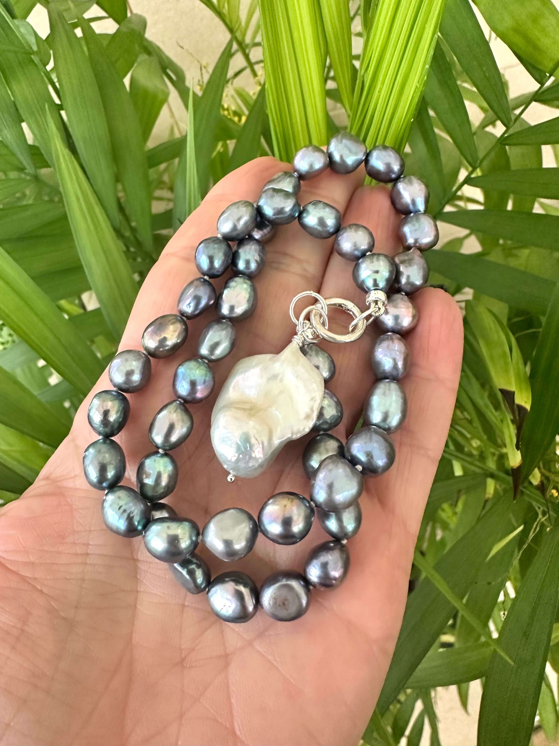 Hand holding black pearl necklace with baroque pendant near green leaf background
