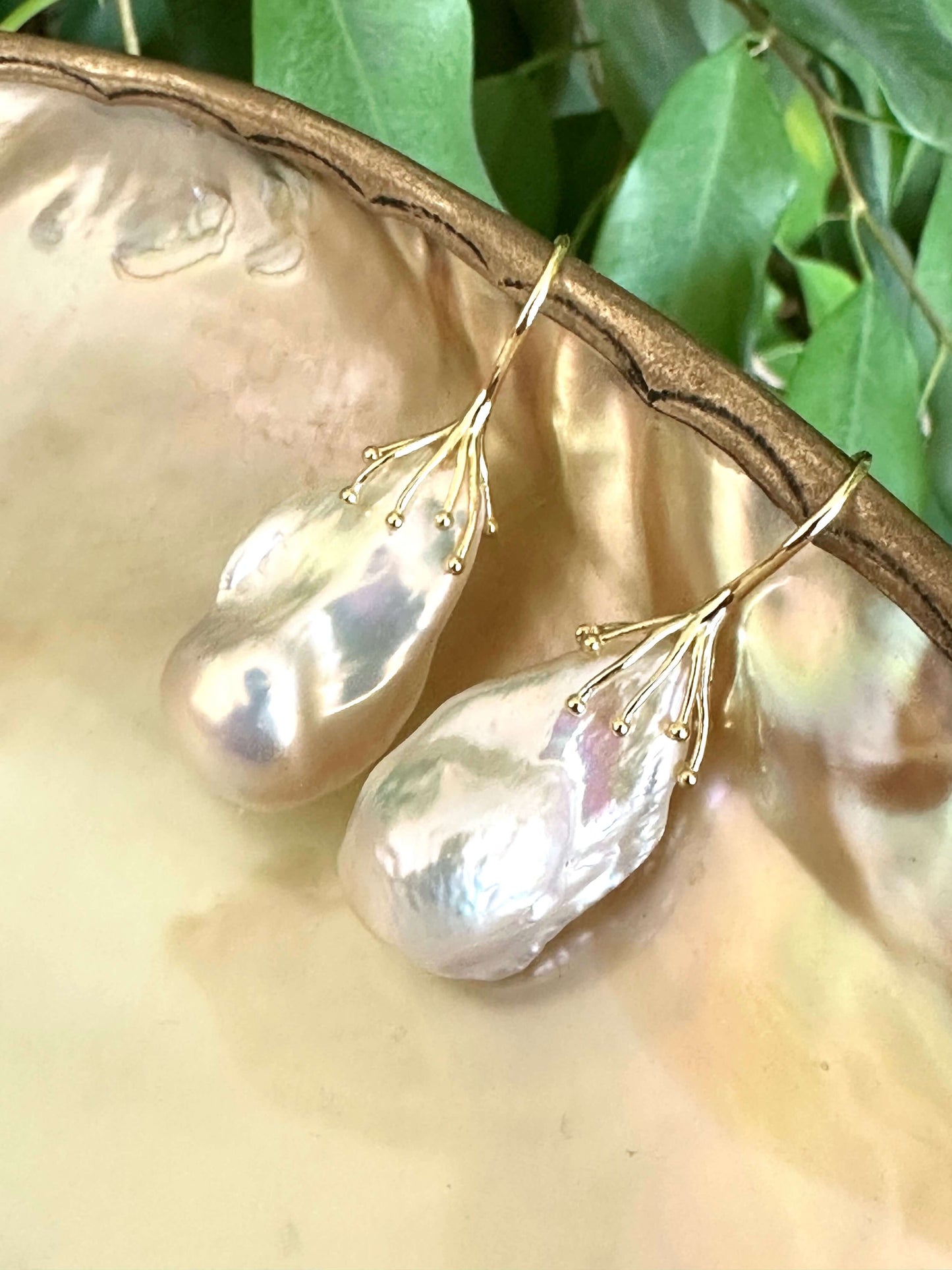 White baroque pearl drop earrings resting on green leaf and pearl shell backdrop, silver hooks visible