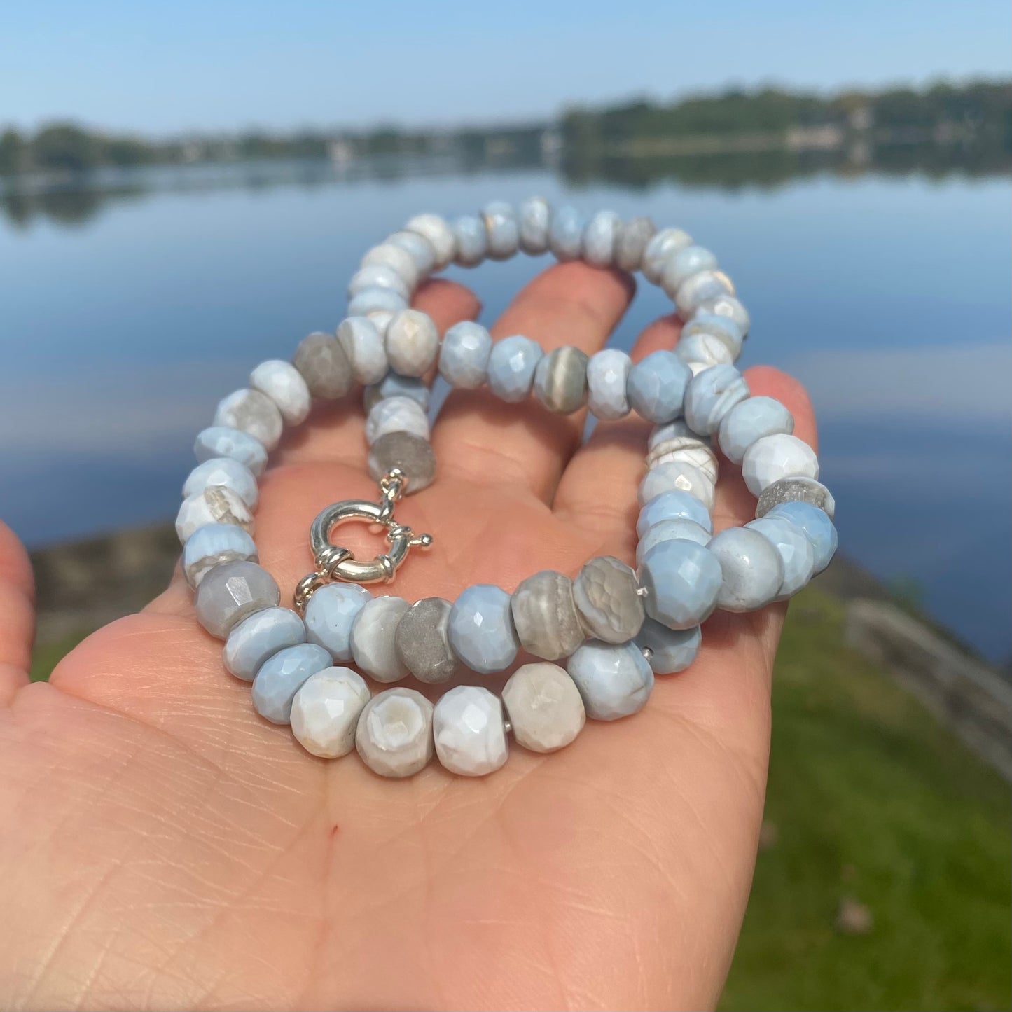 Blue grey opal necklace held in hand near lake and grass backdrop reflecting blue hues