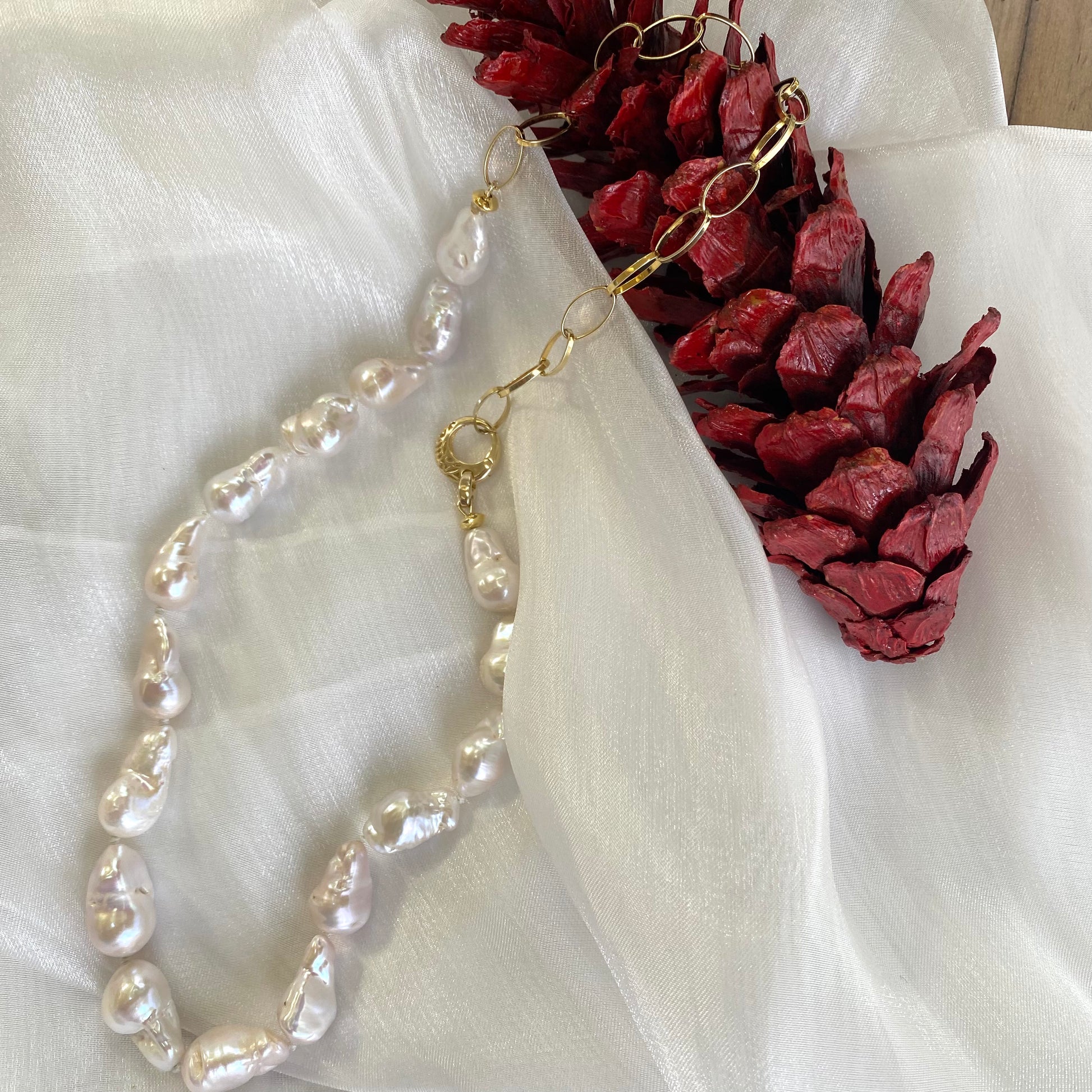 White baroque pearl and gold chain necklace resting on white cloth with red pine cone, elegant handcrafted jewelry from Canada.