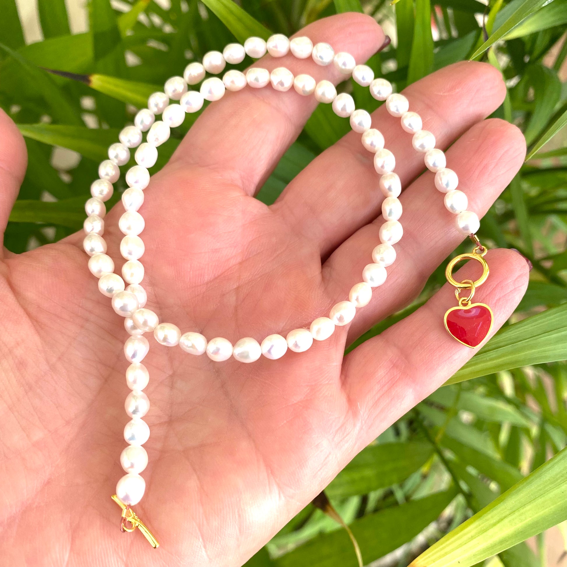 White Pearl Necklace with red heart charm held in hand against green leaf background.