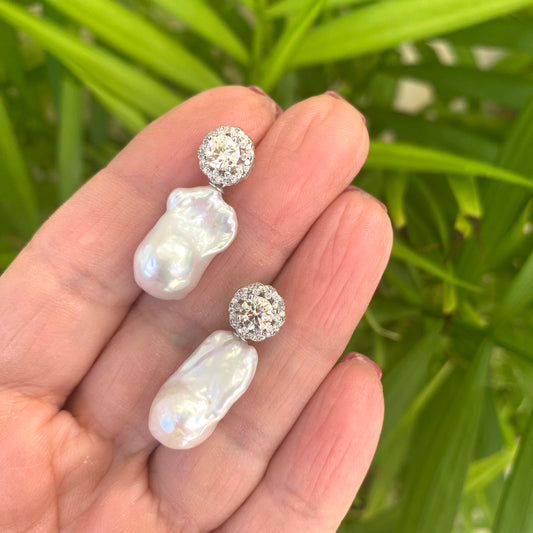 Moissanite and baroque pearl earrings held in hand against green leaves, handmade jewelry display.