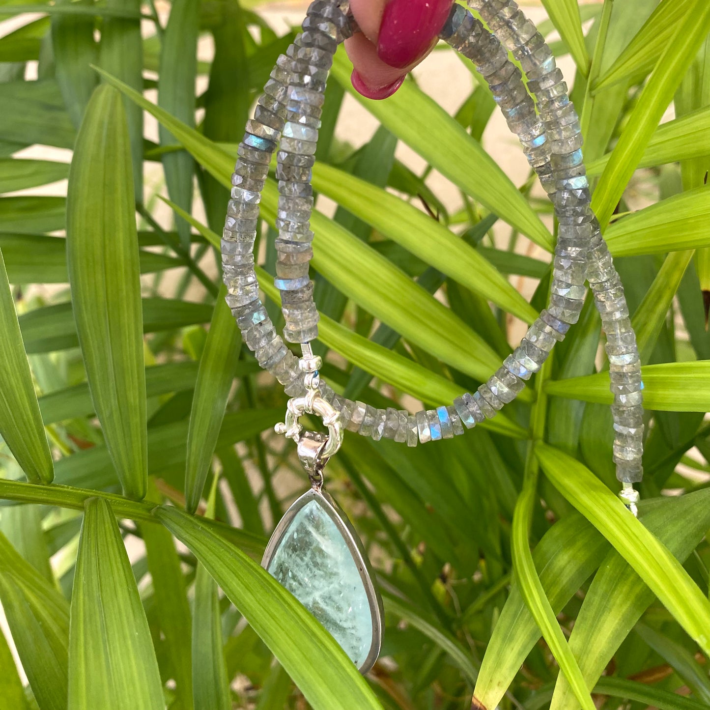 Labradorite aquamarine pendant necklace with aquamarine gemstone held in hand against leafy background — elegant artisan jewelry by Loulia Pearl Jewelry