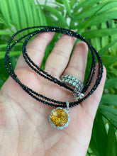 Load image into Gallery viewer, Hand holding black spinel and citrine pendant necklace near green leaf background

