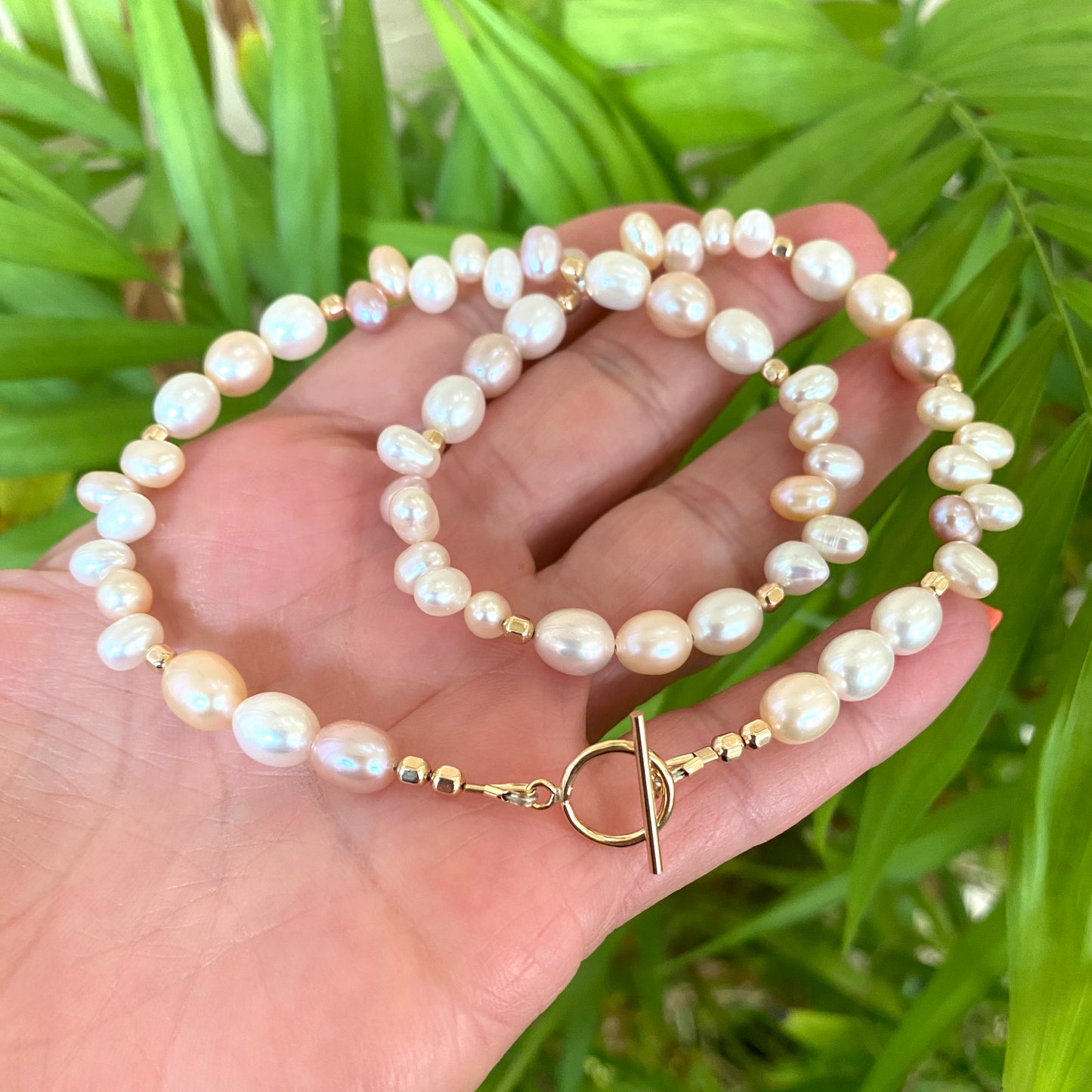 Pearl toggle necklace with pastel pearls and 14K gold filled clasp, held in hand against green backdrop.