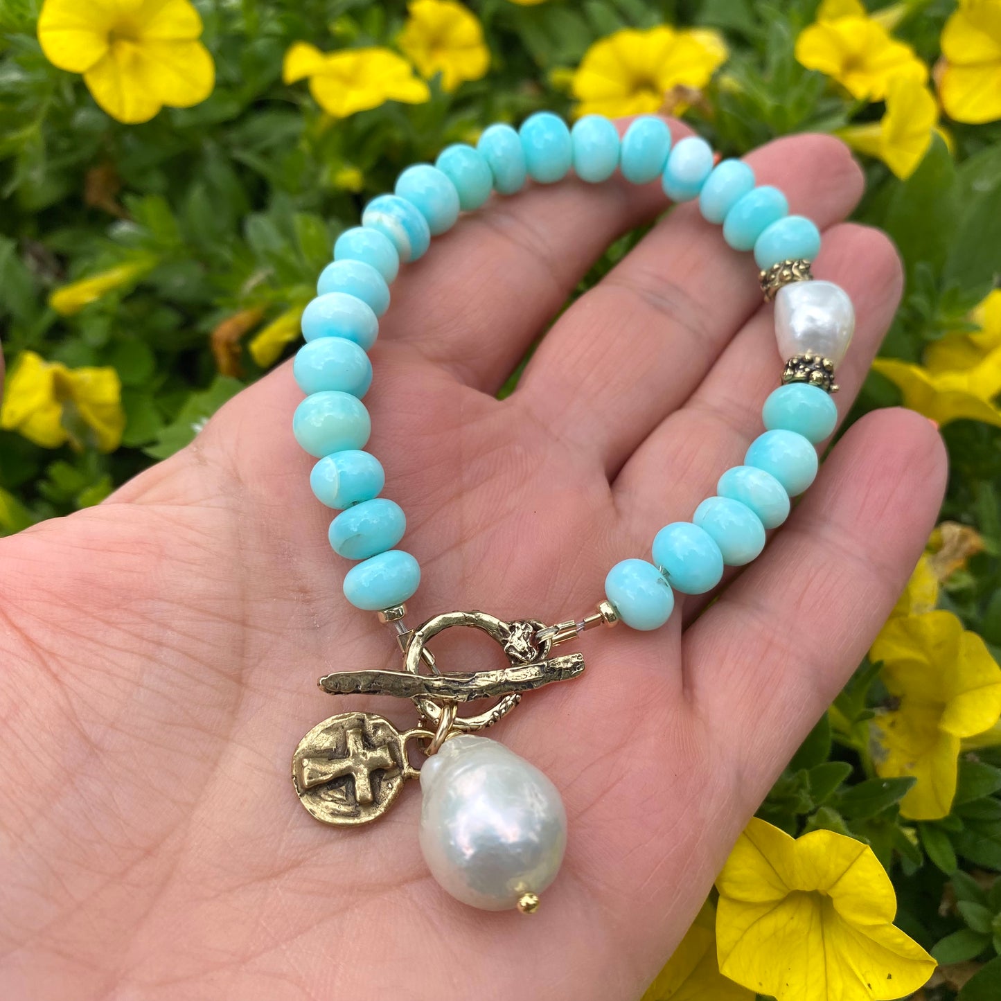 Sky blue opal bracelet with baroque pearls and cross charm showcased in a hand against a backdrop of colorful flowers.