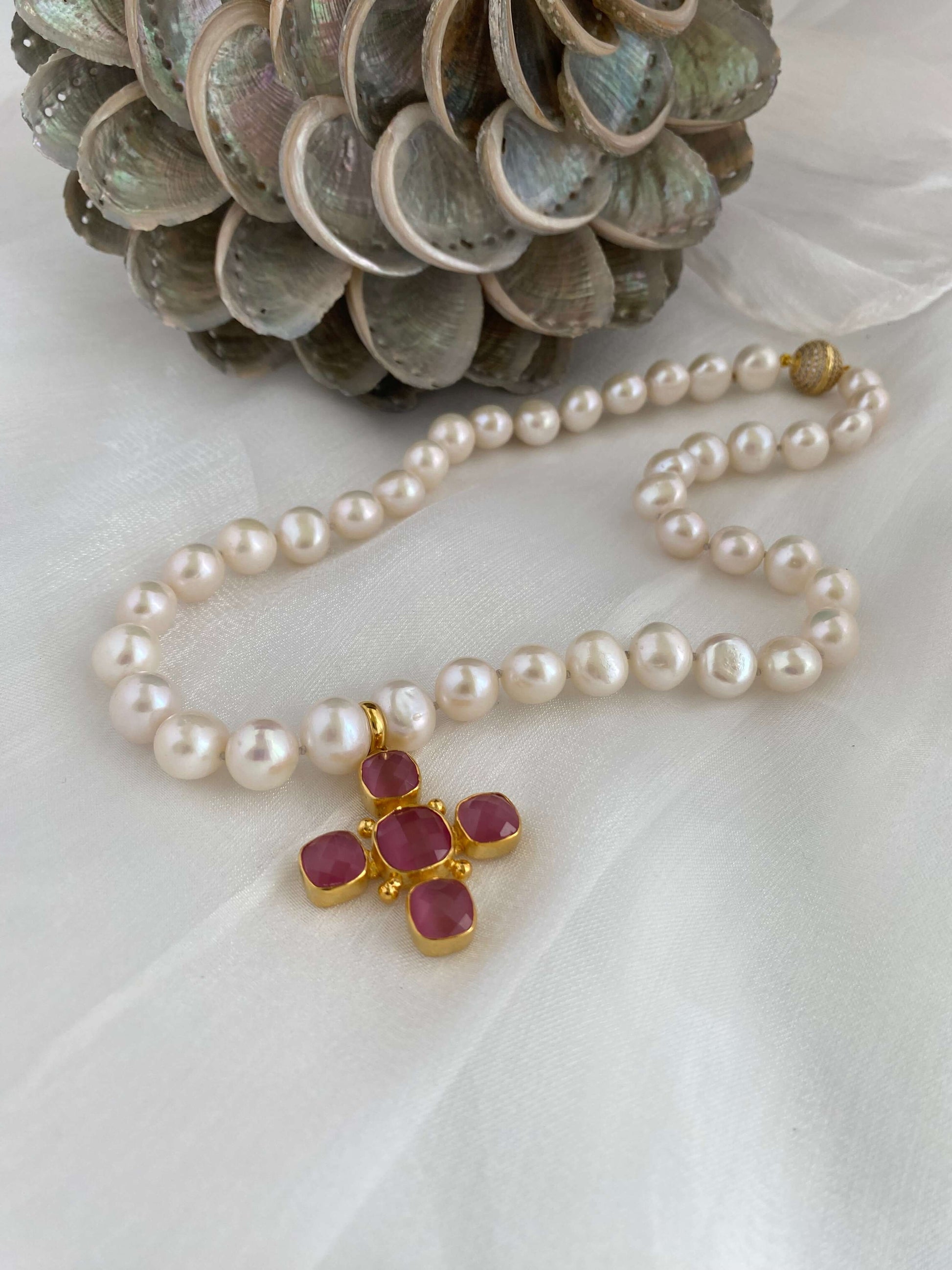 White potato pearl necklace with cross pendant featuring pink accents and a gold clasp, elegantly displayed with a pine cone background.