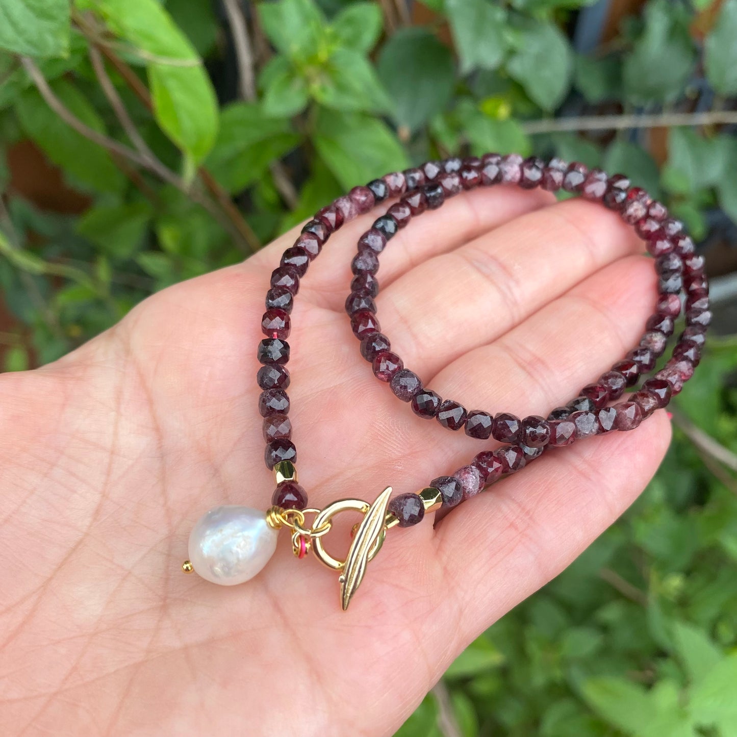 Garnet toggle necklace with baroque pearl charm elegantly displayed in a hand, featuring faceted garnet cube beads.