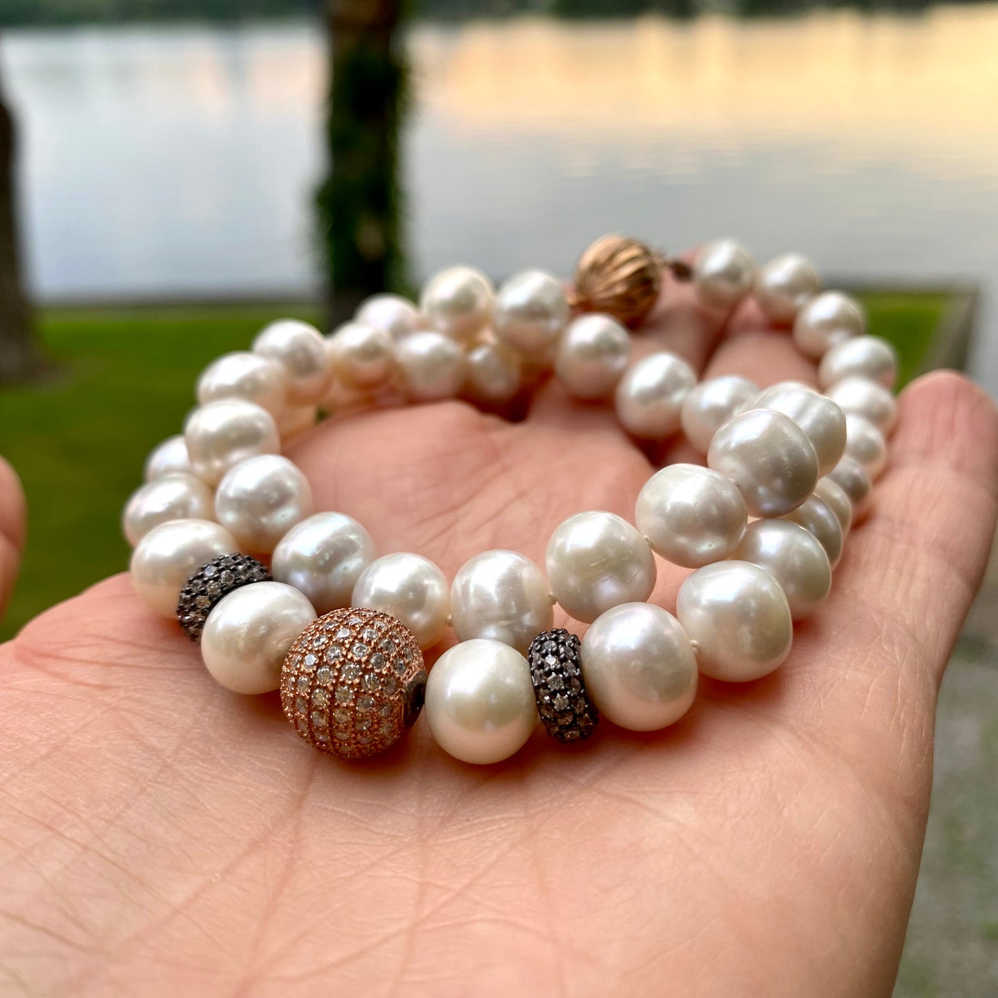 Rose gold pearl necklace held in hand at sunset by a river, showing luminous pearl lustre.