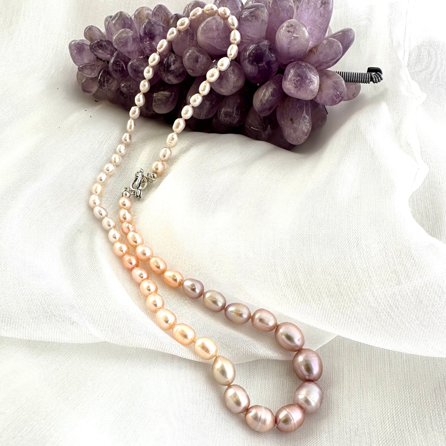 Rice Pearl Necklace with pastel pearls arranged beside amethyst ornament on white fabric background.