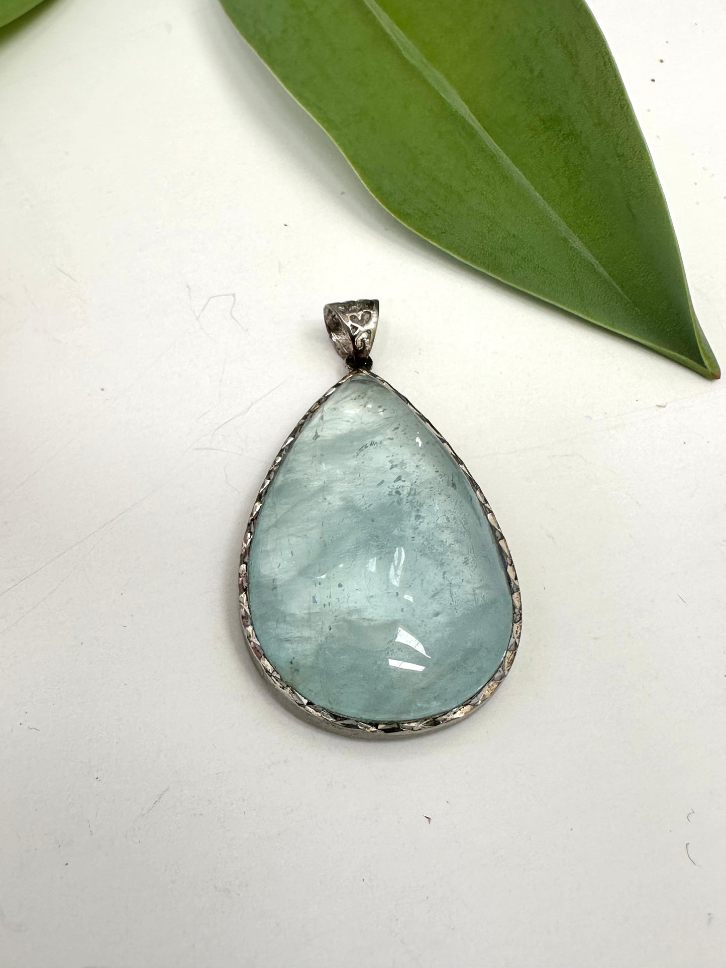 Teardrop-shaped aquamarine pendant on a white background with a green leaf