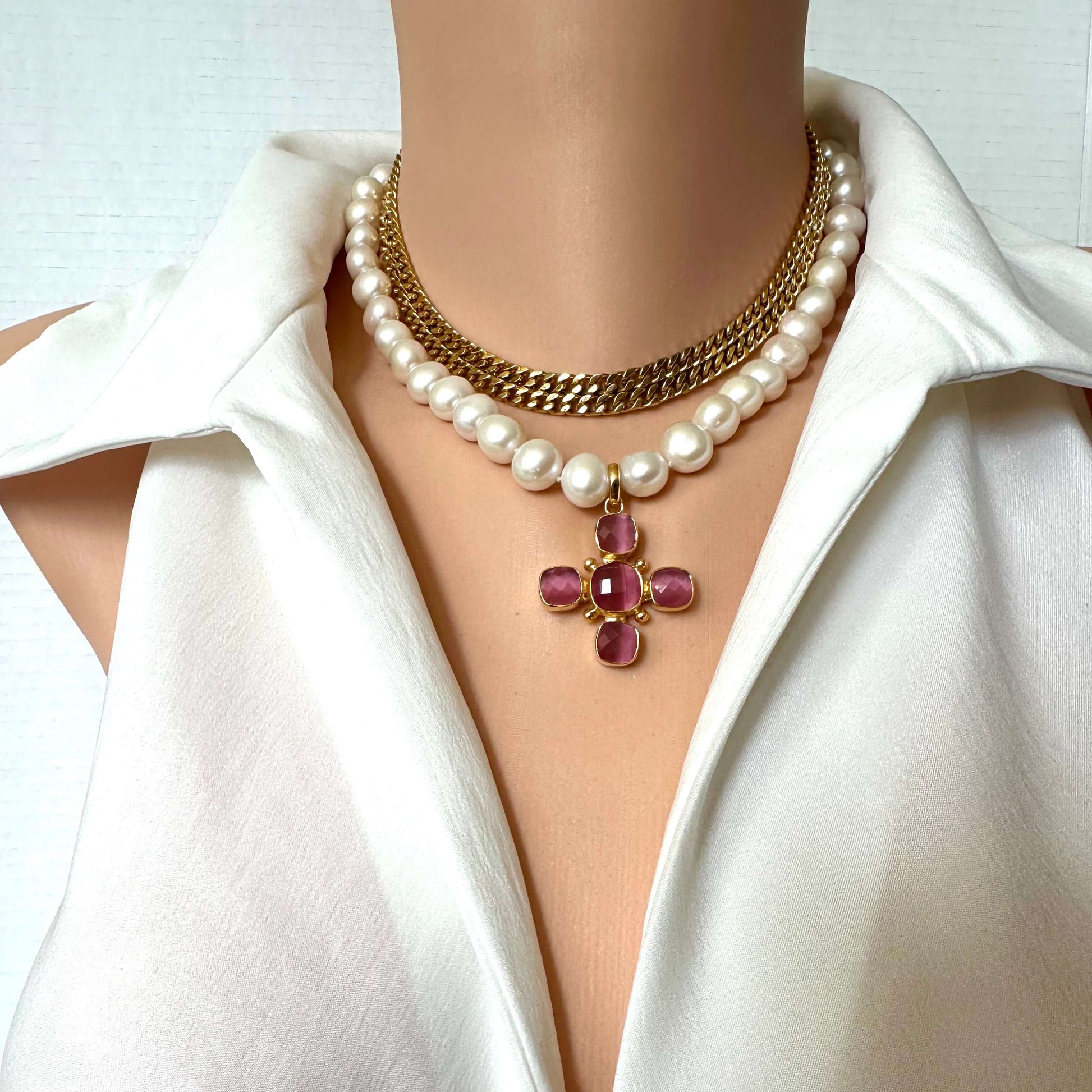 A close-up of a mannequin torso wearing a layered necklace set over a white satin blouse. The necklace features a double-strand pearl strand and a gold chain with a gold cross pendant embellished with pink gemstones.