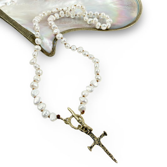 A strand of irregular white freshwater pearls with a small dark red bead, attached to a bronze-toned chain with a decorative dagger pendant, displayed on a white background with a mother-of-pearl shell prop.