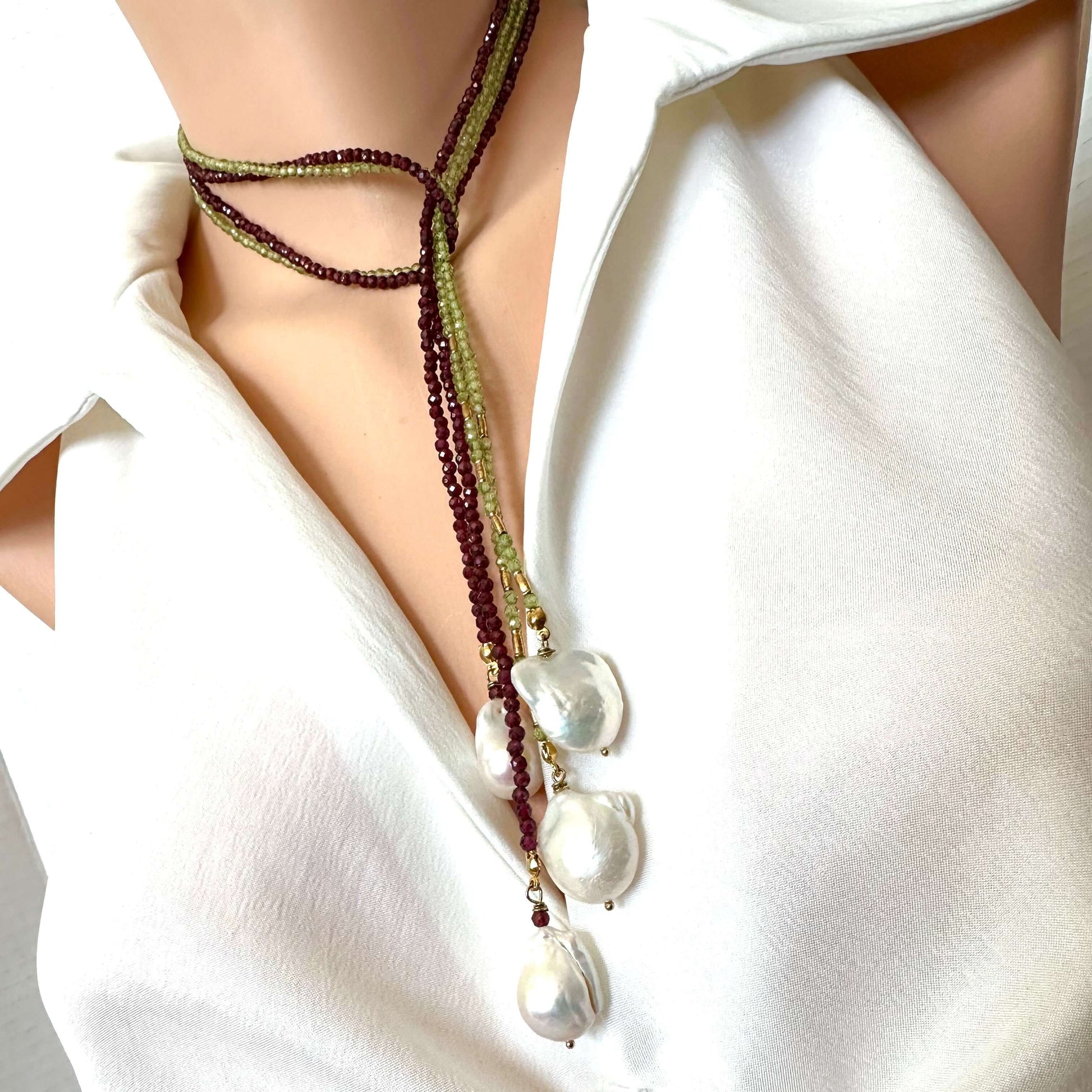 A close-up of a model wearing a multi-strand beaded necklace with large baroque pearls. The necklace features dark purple beads, olive green beads, and gold-toned spacers, styled with a white sleeveless top.