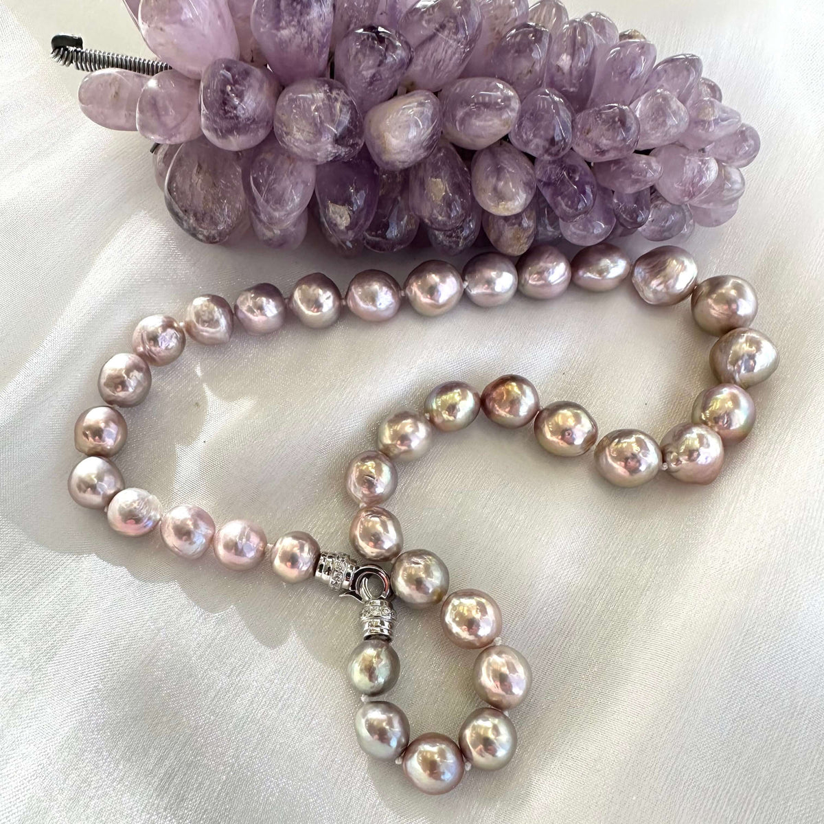 Pink lavender pearl necklace displayed on white cloth beside amethyst crystal ornament, showing elegant color contrast.
