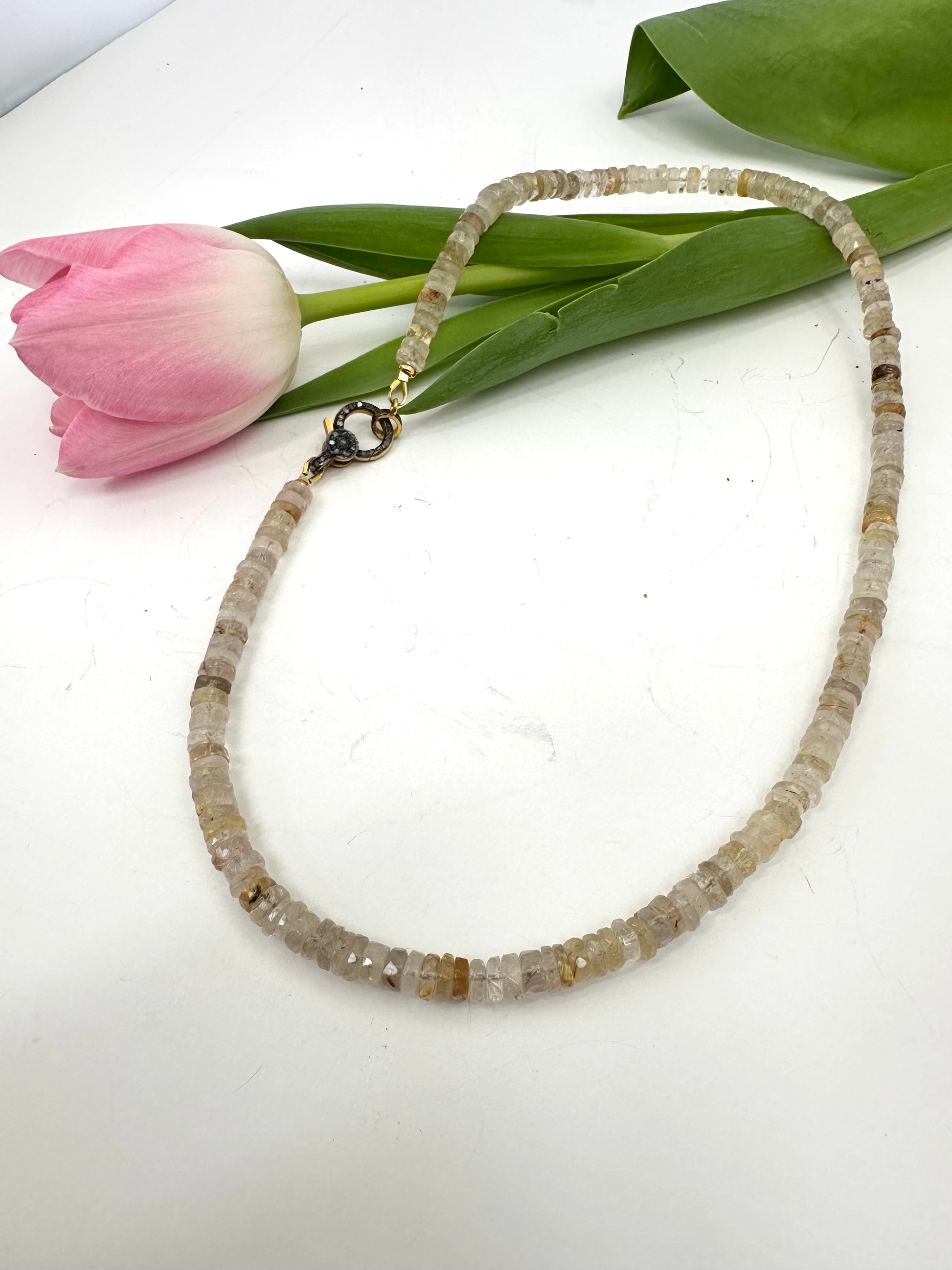 Necklace with beaded chain on a white surface with a pink tulip and green leaves.