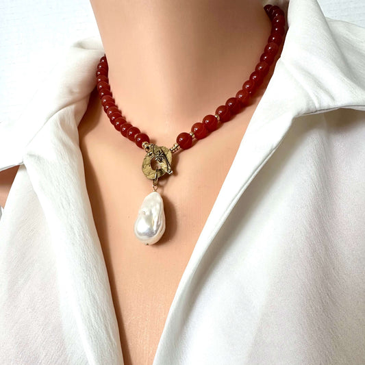 A close-up of a red beaded necklace with a gold-toned decorative clasp and a large white baroque pearl pendant, worn on a mannequin or model with a white collared shirt.