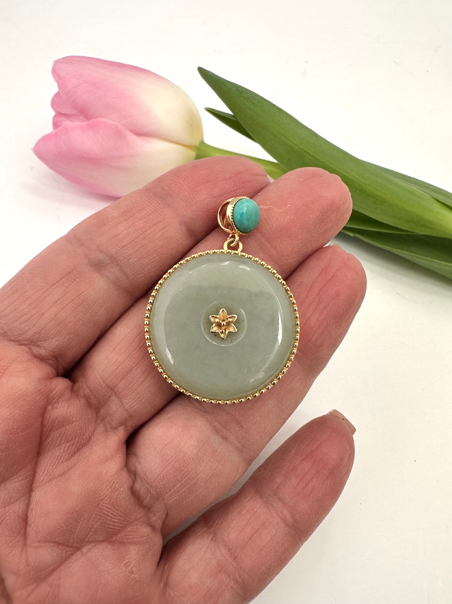 Hand holding a jade pendant with gold accents and a turquoise stone, with pink tulips in the background.