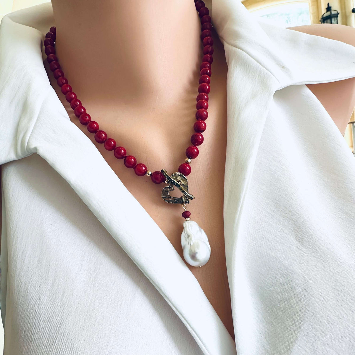 A close-up of a person wearing a red beaded necklace with a dark metal anchor charm and a white baroque pearl pendant, styled with a white collared shirt.