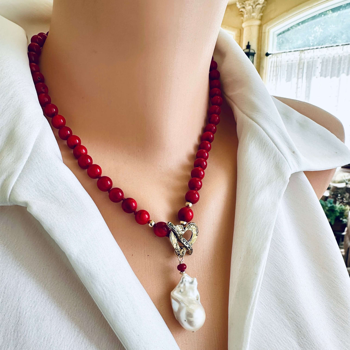 A close-up of a person wearing a red beaded necklace with a silver heart-shaped charm and a white pendant, styled with a white collared shirt.