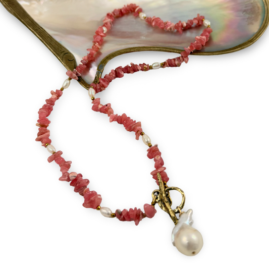 A beaded necklace with chunky coral-pink stone chips, small white pearl-like beads, and a large irregular white baroque pearl pendant, displayed on a white background with a mother-of-pearl fan prop.