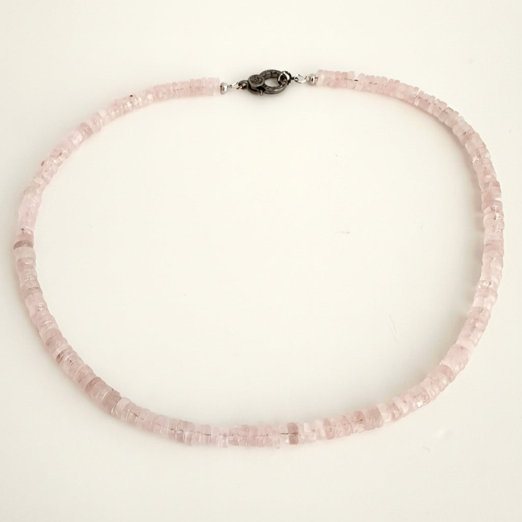 A delicate necklace made of small, faceted light pink beads, featuring a dark metal clasp, displayed on a plain off-white background.