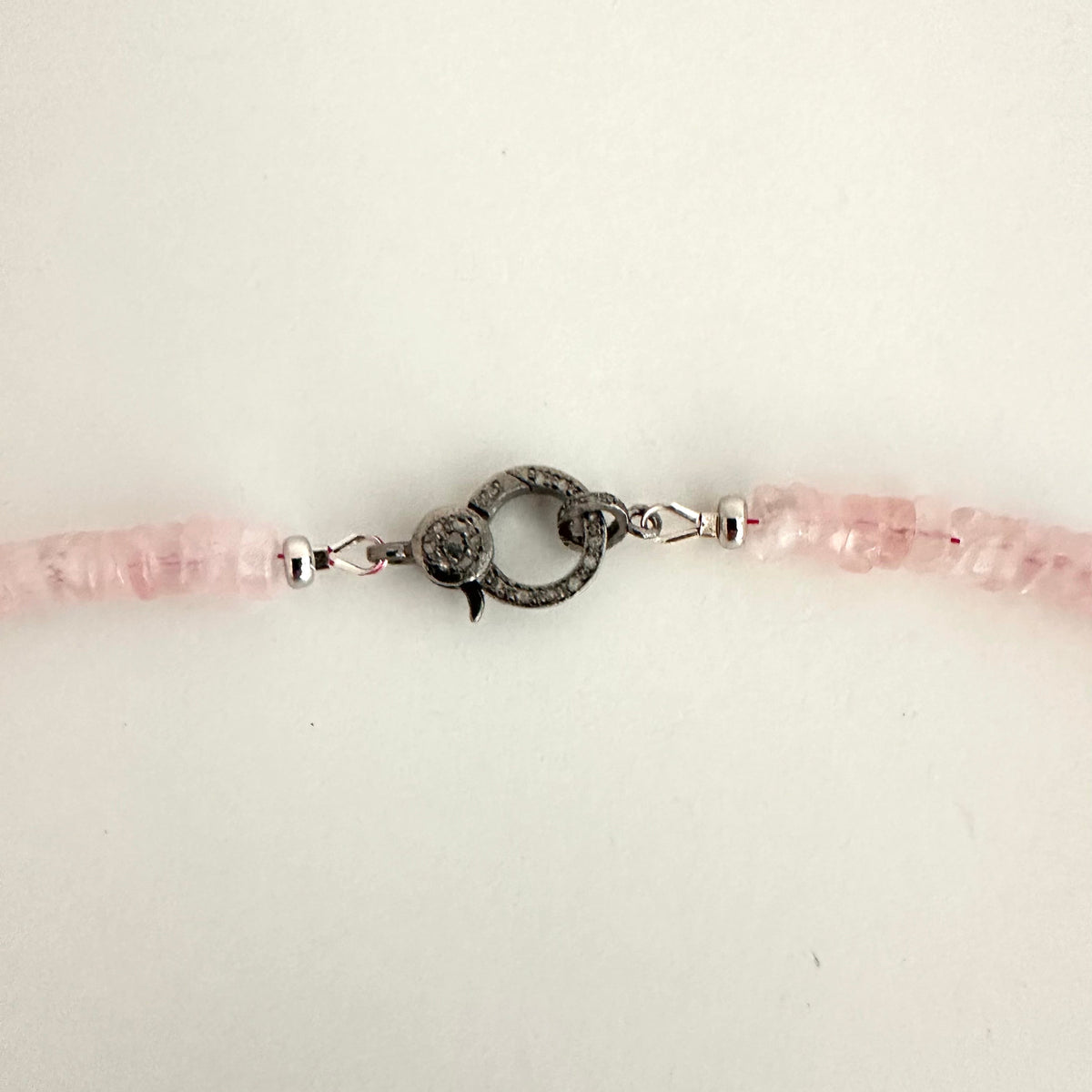 A close-up of a beaded bracelet featuring light pink faceted beads and a central dark gray textured toggle clasp on a plain off-white background.