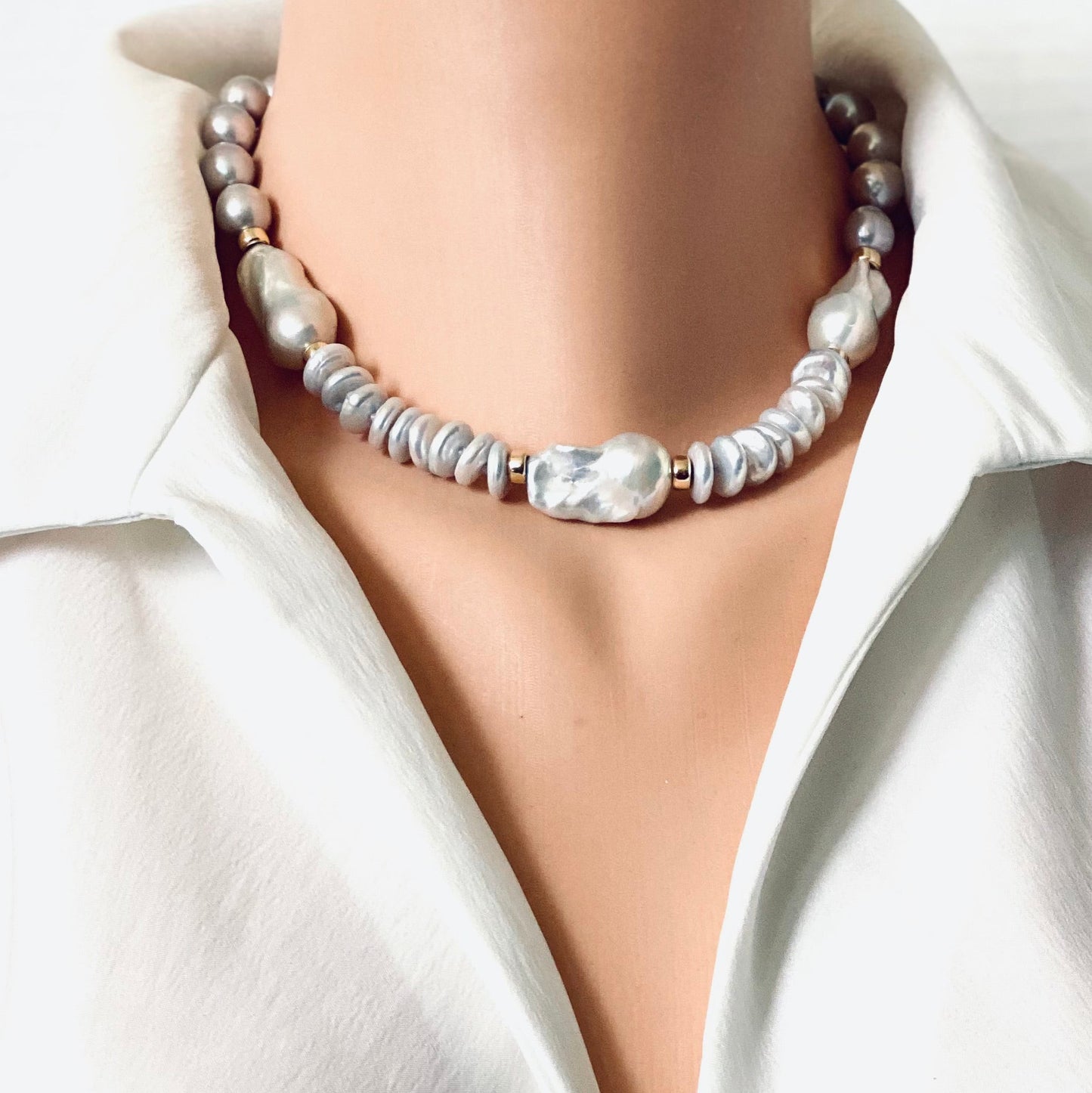 A close-up of a model wearing a pearl necklace with a central textured white BAROQUE and small gold spacer beads, styled with a white satin shirt.