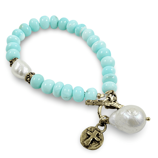 A beaded stretch bracelet featuring light turquoise-blue round beads, a white baroque pearl, antique bronze spacer details, and a dangling cross charm on a white background.